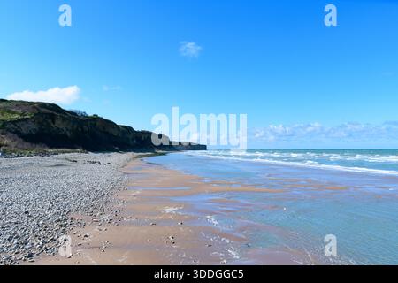 L'ampia vista della spiaggia di Omaha presenta una spiaggia di ciottoli che conduce a scogliere spettacolari, con onde dolci che scivolano sulla sabbia bagnata sotto un cielo blu cristallino. Le texture naturali e la luce del giorno creano un'atmosfera fresca e aperta. Foto Stock