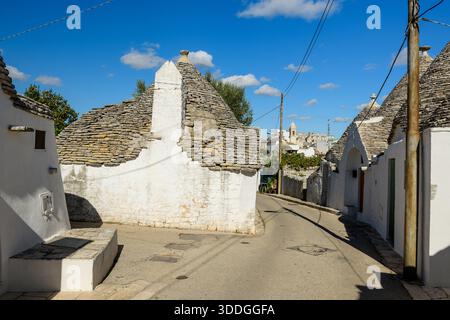 Una strada stretta e tortuosa fiancheggiata da tradizionali case a trullo con tetti conici in pietra e pareti bianche ad Alberobello, in Puglia, in Italia. La scena è illuminata dalla luce solare, evidenziando texture rustiche e forme architettoniche mediterranee contro un cielo blu vivace. Foto Stock