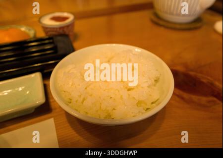 Una ciotola di soffice riso bianco al vapore siede su un tavolo di legno, circondato da eleganti stoviglie giapponesi in un ambiente caldo e invitante per la colazione. L'illuminazione soffusa mette in risalto la consistenza lucida del riso appena cotto. Foto Stock