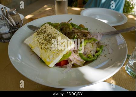 Insalata greca fresca servita in una ciotola bianca, con una fetta spessa di feta, peperoni verdi, cipolle rosse, olive e un filo di olio d'oliva, bagnata dalla luce naturale su un tavolo rustico. Foto Stock