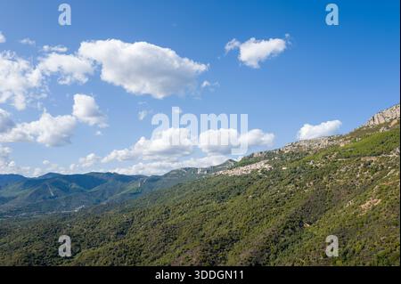Vaste colline ondulate ricoperte di vegetazione lussureggiante si estendono sotto un cielo azzurro punteggiato di nuvole bianche sparse nei pressi di Baunei, in Sardegna. La luce del sole esalta le texture dell'aspro paesaggio mediterraneo. Foto Stock