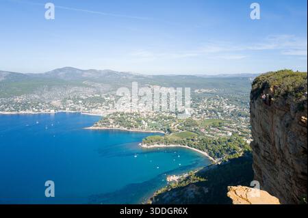 La scogliera rocciosa a strapiombo con vegetazione sparsa si affaccia sulla baia turchese e sulla città costiera di Cassis, con colline ondulate e cielo azzurro cristallino sullo sfondo. La luce del sole mette in evidenza il contrasto tra l'aspra parete rocciosa e le calme acque mediterranee sottostanti. Foto Stock