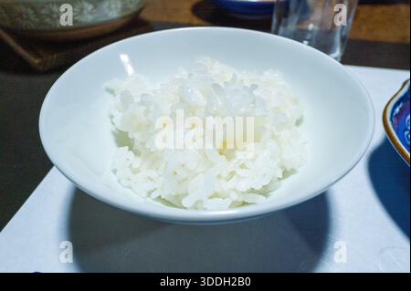 Una porzione di soffice riso bianco al vapore viene presentata in una semplice ciotola in porcellana bianca, che cattura una luce soffusa che mette in risalto la sua consistenza lucida. L'ambiente minimalista evoca un senso di calma e raffinata cucina giapponese. Foto Stock