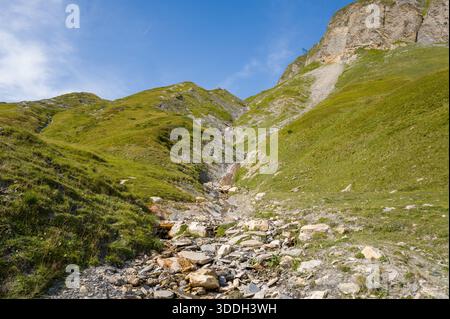 Una piccola cascata si snoda su pietre e ghiaia su una collina verde di montagna sotto aspre scogliere e un cielo azzurro. La luce del sole mette in risalto le fresche consistenze di erba e roccia. Foto Stock