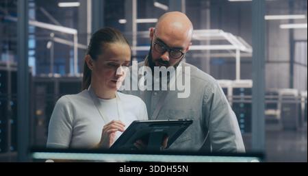 Team di ingegneri IT maschili e femminili che collaborano e discutono dei dati visualizzati su tablet digitali, in piedi in un ambiente sicuro e high-tech dei data center. Concetto Teamwork e Mobile System Diagnostic. Foto Stock