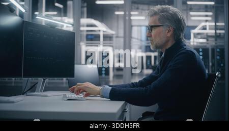 Ingegnere senior specializzato, di mezza età, che digita su tastiera, monitoraggio delle metriche del sistema cloud su due schermi nel data center. Analisi dell'infrastruttura critica e gestione dell'infrastruttura cloud. Foto Stock