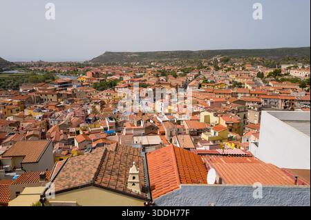 Ampia vista sui tetti in terracotta e sulle facciate pastello di Bosa, in Sardegna, con dolci colline verdi e il fiume in lontananza. La luce soffusa del giorno esalta i toni caldi e l'invitante fascino mediterraneo della città. Foto Stock