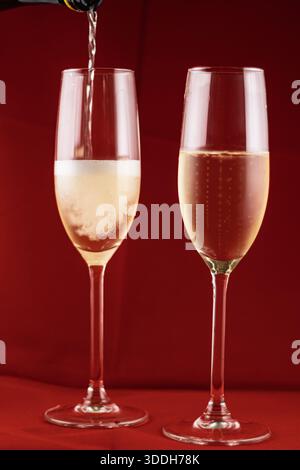 Lo champagne viene versato in due bicchieri per flauto, creando bolle su un vivace sfondo rosso, perfetto per feste e feste Foto Stock