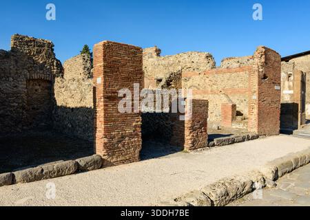 File di antiche colonne in mattoni e muri in pietra intemprata sorgono lungo una strada lastricata nel sito archeologico di Pompei, Italia. La luce solare intensa mette in risalto le consistenze ruvide e le murature conservate sotto un cielo azzurro. Foto Stock