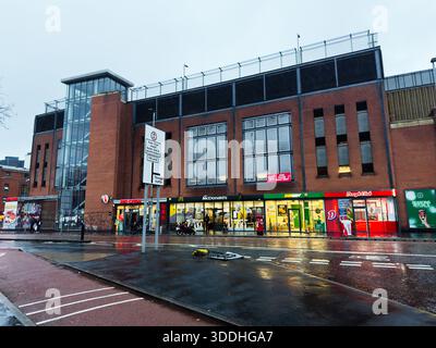 Manchester, Regno Unito - 06 dicembre 2025: Rainy Manchester Street con vetrine in mattoni, torre di vetro e negozi affollati. Foto Stock