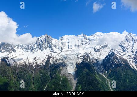Vista mozzafiato del massiccio del Monte bianco, caratterizzato da spettacolari cime innevate, un vasto ghiacciaio e lussureggianti foreste verdi sotto un cielo azzurro vivido con nuvole sparse. La scena trasmette freschezza, scala e grandezza naturale. Foto Stock