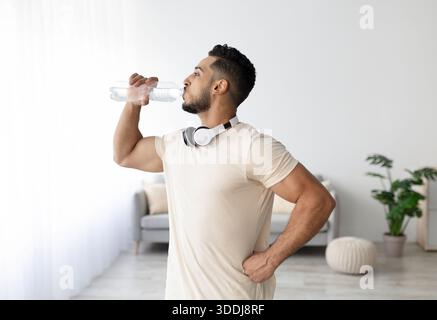 Giovane uomo arabo sportivo con cuffie bere acqua dopo il suo allenamento a casa Foto Stock