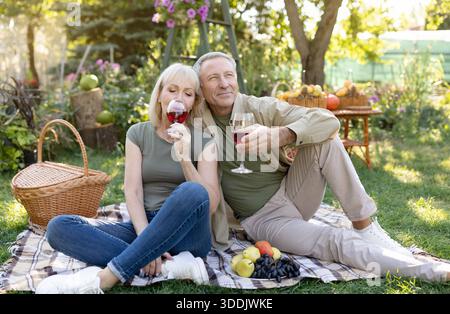 I coniugi anziani eccitati festeggiano l'anniversario, bevono vino, si siedono sulla coperta mentre fanno un picnic nel loro giardino Foto Stock