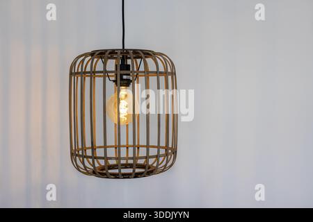 Lampada a sospensione decorativa con lampadina incandescente in una gabbia in tessuto naturale contro le pareti bianche Foto Stock
