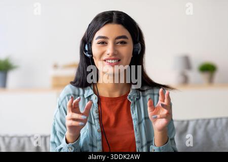 Allegra donna araba che effettua videochiamate da casa indossando le cuffie durante le riunioni online Foto Stock