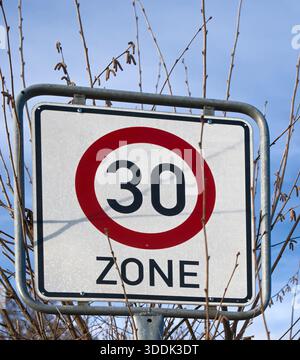 Un segnale stradale di 30 km/h si trova su uno sfondo di rami di alberi e cielo, che indica una zona limite di velocità in un'area suburbana Foto Stock