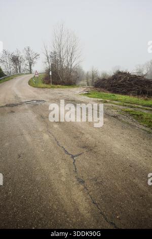 Bivio tra una strada su un terrapieno, che conduce ad un ponte, e un sentiero fangoso, che conduce ad un fiume incorniciato da alberi, in una giornata nebbiosa e nuvolosa nell'ita Foto Stock