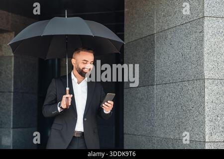 Uomo d'affari felice e maturo che usa lo smartphone tenendo in mano l'ombrello nero durante la passeggiata in città il giorno della pioggia Foto Stock