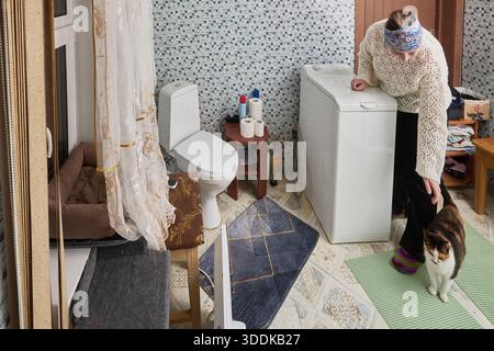 L'impostazione del programma di lavaggio sulla lavatrice con caricamento dall'alto la donna di circa 50 anni viene distratta dal gatto. Foto Stock