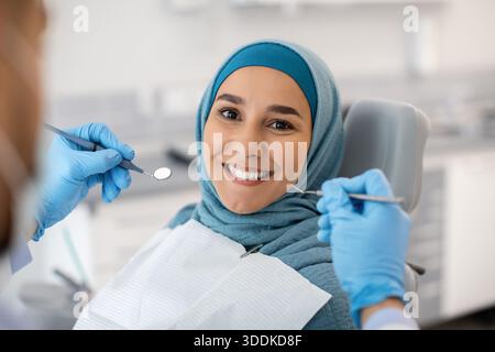 Donna musulmana felice seduta alla sedia dentista e sorridente alla camera Foto Stock