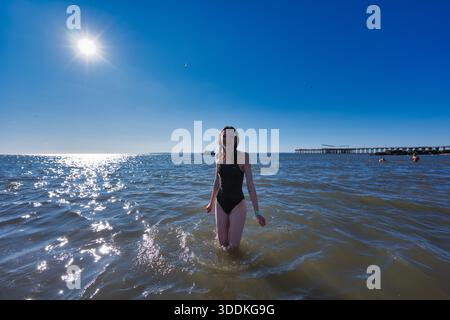 Una giovane donna con un costume da bagno nero si erge nell'oceano poco profondo in una giornata luminosa e soleggiata. Il sole si riflette brillantemente sull'acqua. Foto Stock