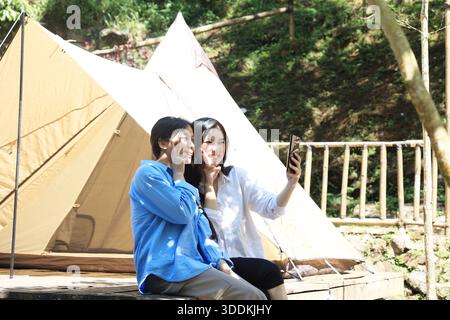 Due donne asiatiche fanno un selfie fuori da una tenda, la giovane donna sorride calorosamente con V Peace pose Foto Stock