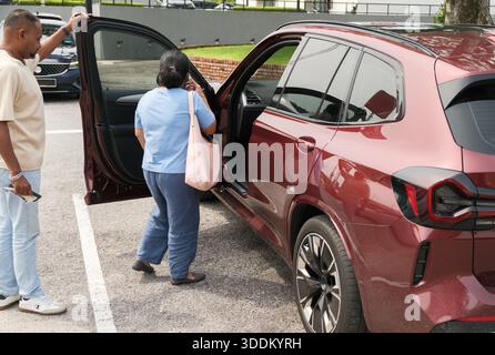 Una donna sta entrando in macchina mentre un uomo sta fuori tenendo un telefono. Si trovano in un'area di parcheggio con altri veicoli intorno a loro. Il sole è shini Foto Stock