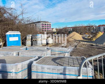 Un cantiere con pali di sabbia e ghiaia e pallet di pietre per pavimentazione avvolti in pellicola di plastica. Una recinzione metallica è visibile in primo piano, wh Foto Stock