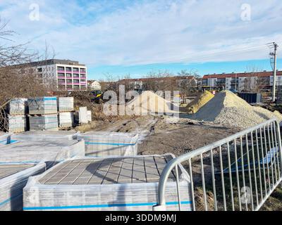 Un cantiere con pali di sabbia e ghiaia e pallet di pietre per pavimentazione avvolti in pellicola di plastica. Una recinzione metallica è visibile in primo piano, wh Foto Stock