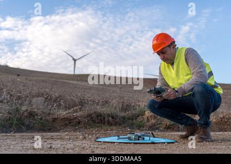 Ingegnere che indossa giubbotto di sicurezza che utilizza un telecomando con drone su un piazzale di atterraggio, preparandosi per l'ispezione delle turbine eoliche Foto Stock