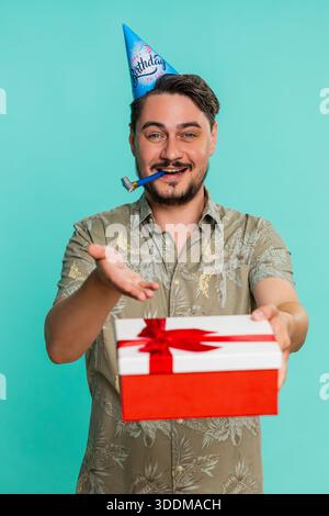 Un uomo di mezza età che indossa un cappello festoso sorride calorosamente, fa felicemente un regalo di compleanno, celebrando chiaramente la generosità e la gentilezza. Ragazzo adulto isolato su sfondo blu che si gode una festa gioiosa Foto Stock