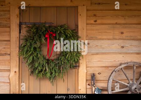 Tradizionale corona natalizia fatta di rami sempreverdi appesi su una porta rustica in legno di una capanna di legno. Corona di abete verde naturale con nastro rosso. Foto Stock