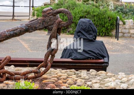 Persona seduta da sola su una panchina di legno con indosso una giacca nera con cappuccio accanto a una grande nave arrugginita ancorata sul lungomare di Ventnor, Isola di wight, Regno Unito Foto Stock
