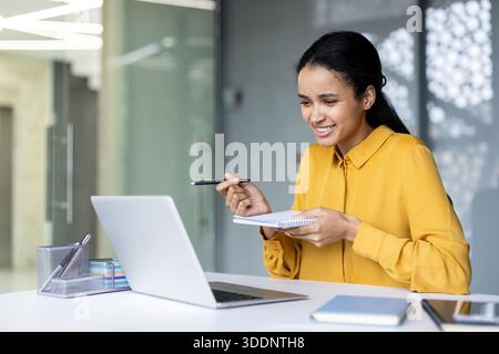 Giovane donna con una camicia gialla sorridente e attiva che partecipa a una riunione virtuale o a una lezione online, prendendo appunti con una penna e guardando un computer portatile Foto Stock