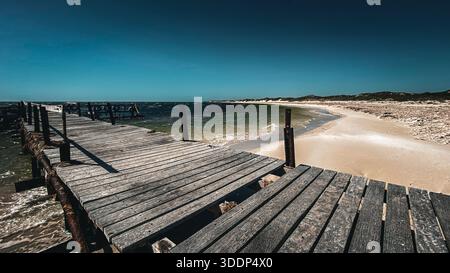 Un molo di legno intemprato si estende nell'oceano blu, circondato da una spiaggia tranquilla. Questa scena evoca tranquillità e tranquillità. Foto Stock