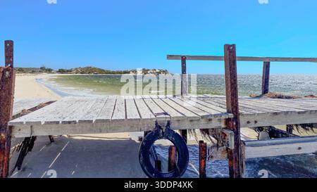Un molo di legno intemprato si estende su una spiaggia sabbiosa, in contrasto con il sereno oceano e il limpido cielo blu, evocando tranquillità. Foto Stock