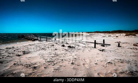 Un molo di legno intemprato si estende nell'oceano instabile, circondato da spiaggia sabbiosa e cielo limpido, evocando tranquillità e avventura. Foto Stock