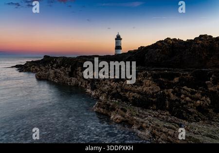 Faro di Penmon Point all'alba sopra una costa rocciosa e calme acque costiere Foto Stock