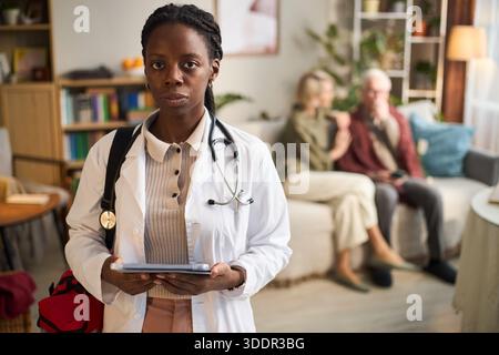 Ritratto di giovane donna nera adulta medico in piedi con tablet digitale, con stetoscopio, guardando la macchina fotografica, anziana coppia caucasica seduta sul divano sullo sfondo, ambientazione di visita a casa Foto Stock