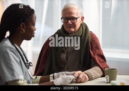 Uomo caucasico anziano seduto a tavola tenendosi per mano con giovane infermiera donna nera adulta che indossa stetoscopio, avendo lo zucchero nel sangue testato, entrambi impegnati in conversazione in ambiente sanitario Foto Stock