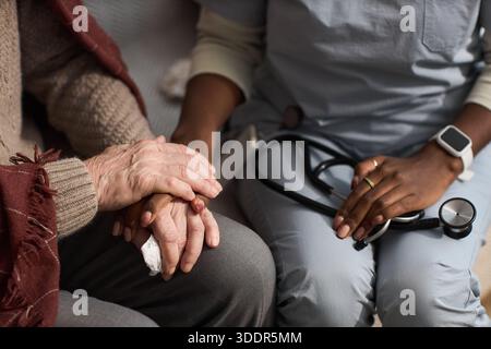 Senior uomo caucasico seduto con le mani appoggiate in grembo, essere confortato da giovane donna nera adulta che tiene lo stetoscopio, mostrando sostegno e cura in ambiente medico Foto Stock