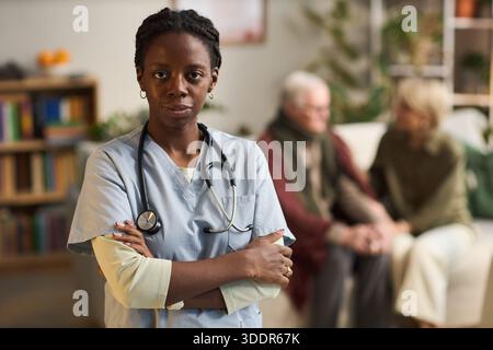 Ritratto di giovane adulto donna nera infermiera in piedi con le braccia incrociate, indossando scrub medici e stetoscopio, anziana coppia caucasica seduta insieme sullo sfondo Foto Stock