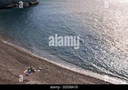 donna che prende il sole su una coperta rossa su una spiaggia di ciottoli vulcanici vicino al mare blu con la scintillante luce del sole Foto Stock