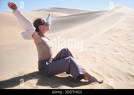 Giovane donna seduta su morbide dune di sabbia con le braccia alzate, godendo del paesaggio desertico aperto sotto la luce del sole. Libertà, gioia, consapevolezza, viaggi. Sabbia Foto Stock