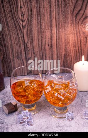 Primo piano di due bicchieri da cecchino di brandy riempiti di whisky e cubetti di ghiaccio sulla superficie ruvida, sfondo caldo a lume di candela. Bar e concetto di festa Foto Stock