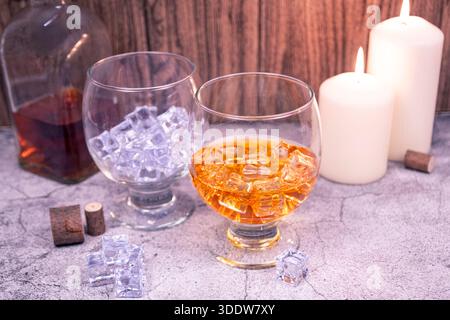 Due cecchini su un tavolo di pietra: Uno con whisky e ghiaccio, uno con ghiaccio. Bottiglia e candele creano un ambiente accogliente. Foto Stock