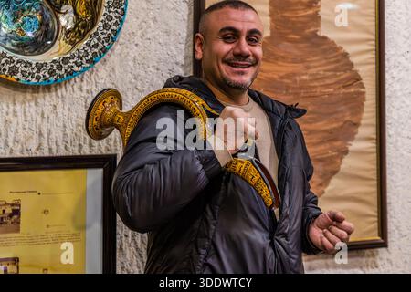 Un dimostrante presso Chez Galip Pottery (Çeç Pottery) presenta un’anfora dipinta artisticamente ad Avanos, in Turchia. Porta il vaso a tracolla come un trofeo. Uno piatto di ceramica dipinto è appeso in secondo piano accanto a un grande murale di un vaso di argilla. Dal 1979, la bottega gestisce anche il museo adiacente dei Capelli, dove sono appese ciocche di più di 13.000 donne. Foto Stock