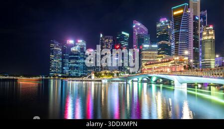 Vivace vista notturna dello skyline di Marina Bay di Singapore con grattacieli illuminati e un ponte affollato che si riflette sull'acqua calma. Questa immagine del paesaggio urbano con Foto Stock