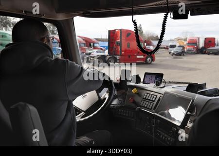 Il conducente si siede in una cabina del veicolo, girando la ruota mentre è parcheggiato in un parcheggio pieno di diversi veicoli. Il tempo è nuvoloso e si vedono diversi camion Foto Stock