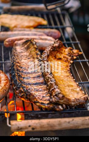 Primo piano verticale di due costolette di maiale dorate che arrostono lentamente su una griglia per barbecue con salsicce e filetti di pollo sullo sfondo. Foto Stock